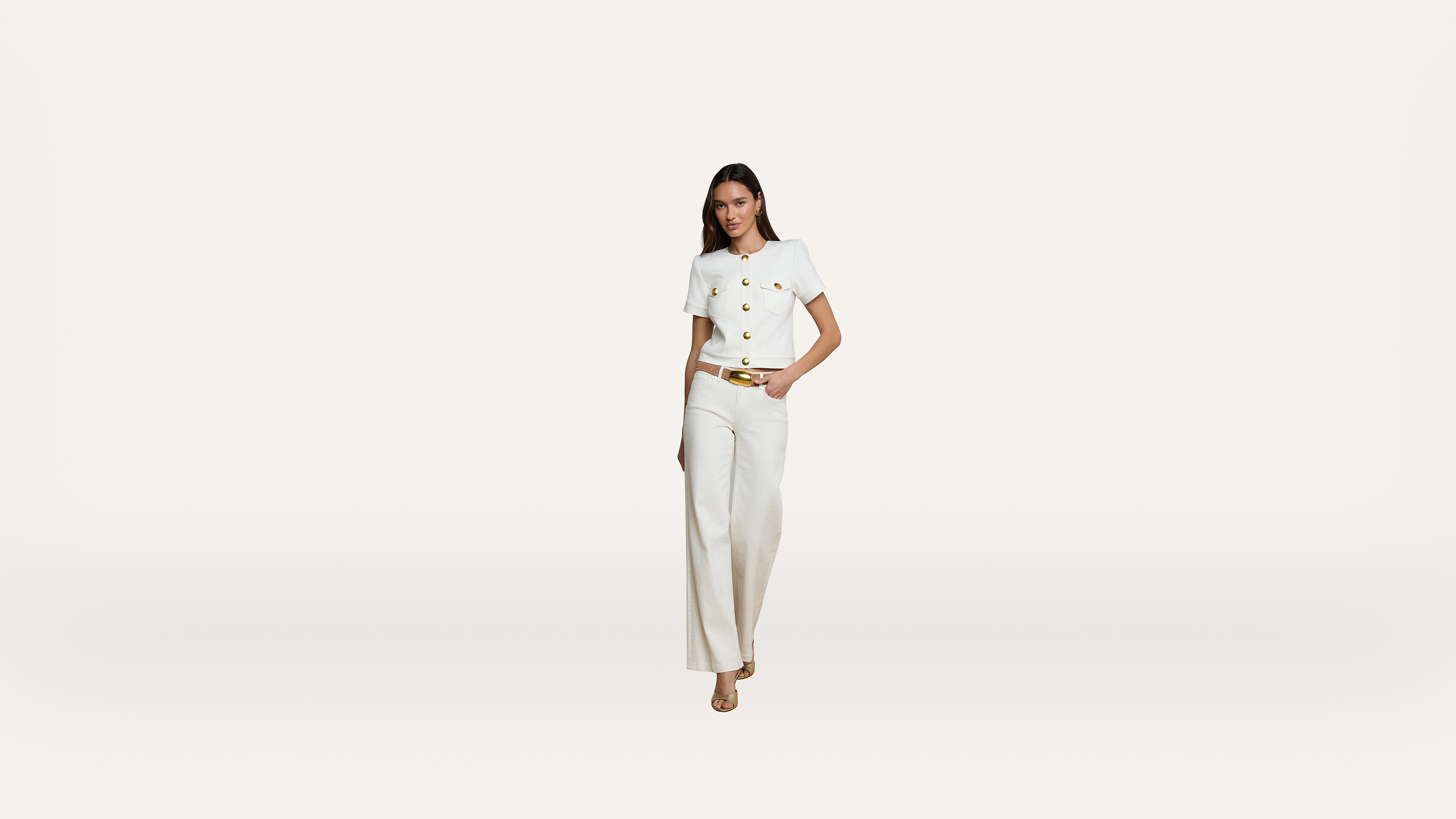 A woman wearing a white short-sleeved top with gold buttons and white high-waisted trousers stands against a plain white background. She has one hand in her pocket and is wearing beige heels.