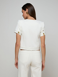A woman stands with her back to the camera, wearing the Nawa Shirt Jacket—a tailored, collarless white piece with gold buttons on the sleeves—paired with matching white pants. Her straight brown hair and gold earrings complement the ensemble against a plain light background.