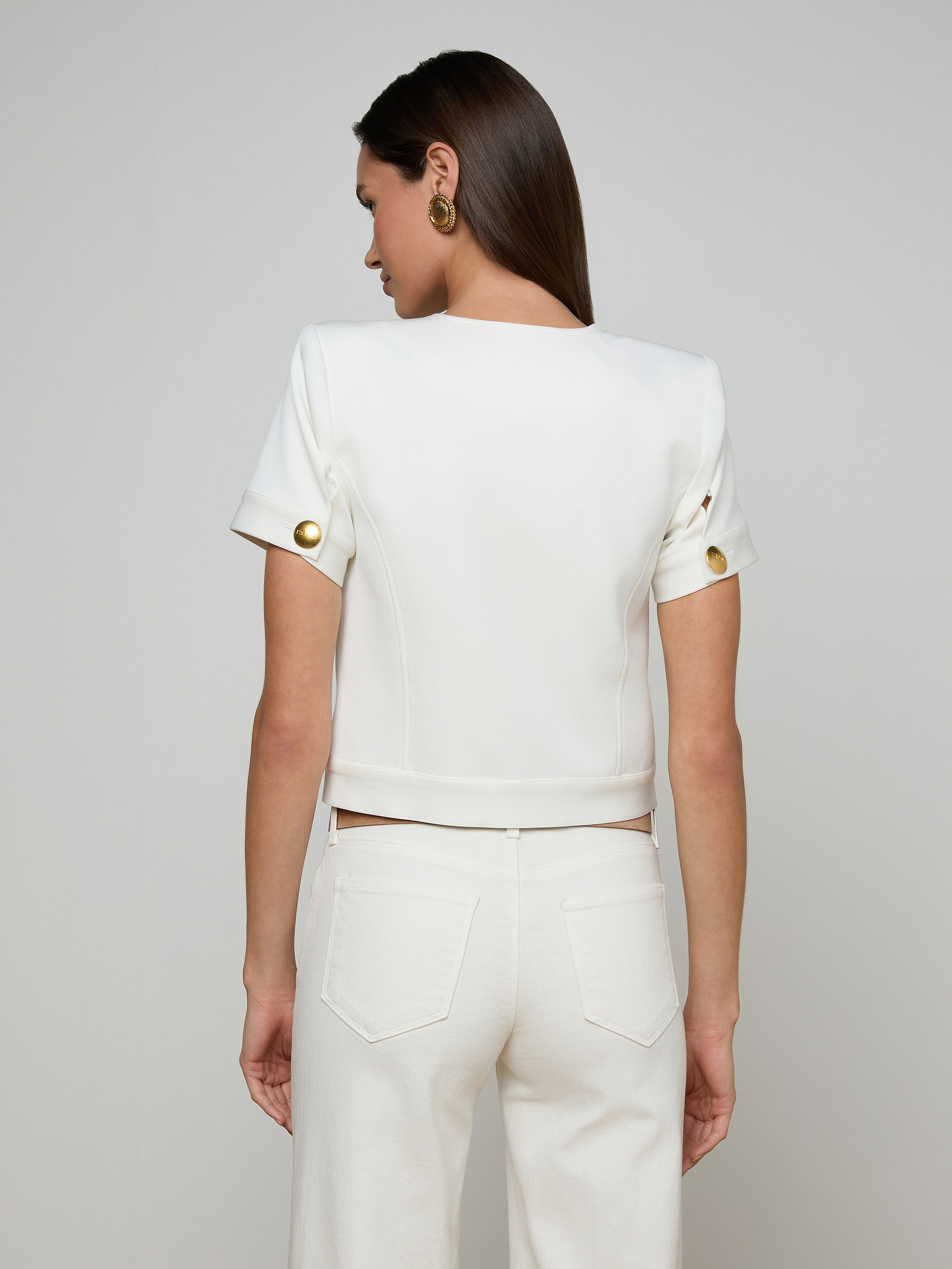 A woman stands with her back to the camera, wearing the Nawa Shirt Jacket—a tailored, collarless white piece with gold buttons on the sleeves—paired with matching white pants. Her straight brown hair and gold earrings complement the ensemble against a plain light background.