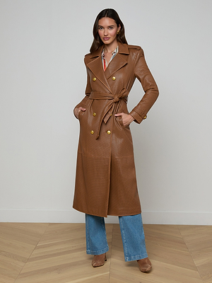 A woman stands indoors in the Baylee Leather Trench Coat—a long, brown lambskin belted trench with gold buttons—styled with blue jeans, tan heeled boots, and a patterned shirt. She smiles slightly at the camera, hands in her pockets.