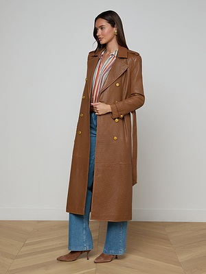 A woman stands indoors wearing the Baylee Leather Trench Coat over a striped blouse, blue jeans, and brown heels. She has long hair and gold earrings, on a light wooden floor against a plain wall.