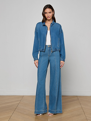 A woman with long, wavy hair stands indoors on a wooden floor, wearing the Dunia Jean Jacket, wide-leg high-waisted jeans, a white top, and sandals. She looks calm against a plain light-colored wall.