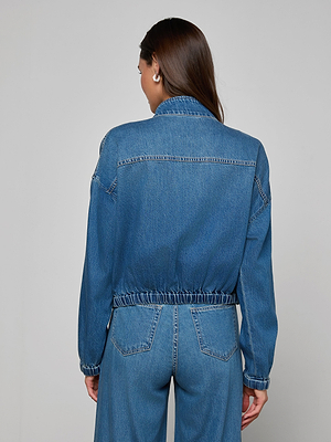 A woman with long brown hair, seen from behind, wears the Dunia Jean Jacket and blue denim jeans against a plain light gray background.