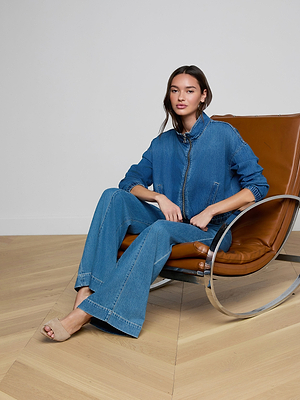 A woman sits in a tan leather chair on a wooden floor, wearing the Dunia Jean Jacket with matching wide-leg jeans and beige heeled mules. The background is plain and light-colored.