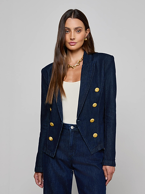 A woman with long brown hair wears the L'AGENCE Emerson Blazer, a double-breasted denim jacket with large gold buttons, over a cream top and matching jeans. She accessorizes with gold earrings and a chunky gold necklace.