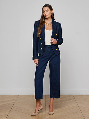 A woman stands indoors wearing the Emerson Blazer by L'AGENCE, a dark double-breasted denim style with gold buttons, paired with matching pants, a white top, beige heels, long brown hair, and a gold necklace.