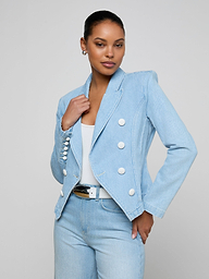 A woman stands confidently against a plain light background, wearing the Emerson Structured Blazer—a light wash, all-cotton denim double-breasted blazer with white buttons—paired with a white top, light blue jeans, and a black belt.