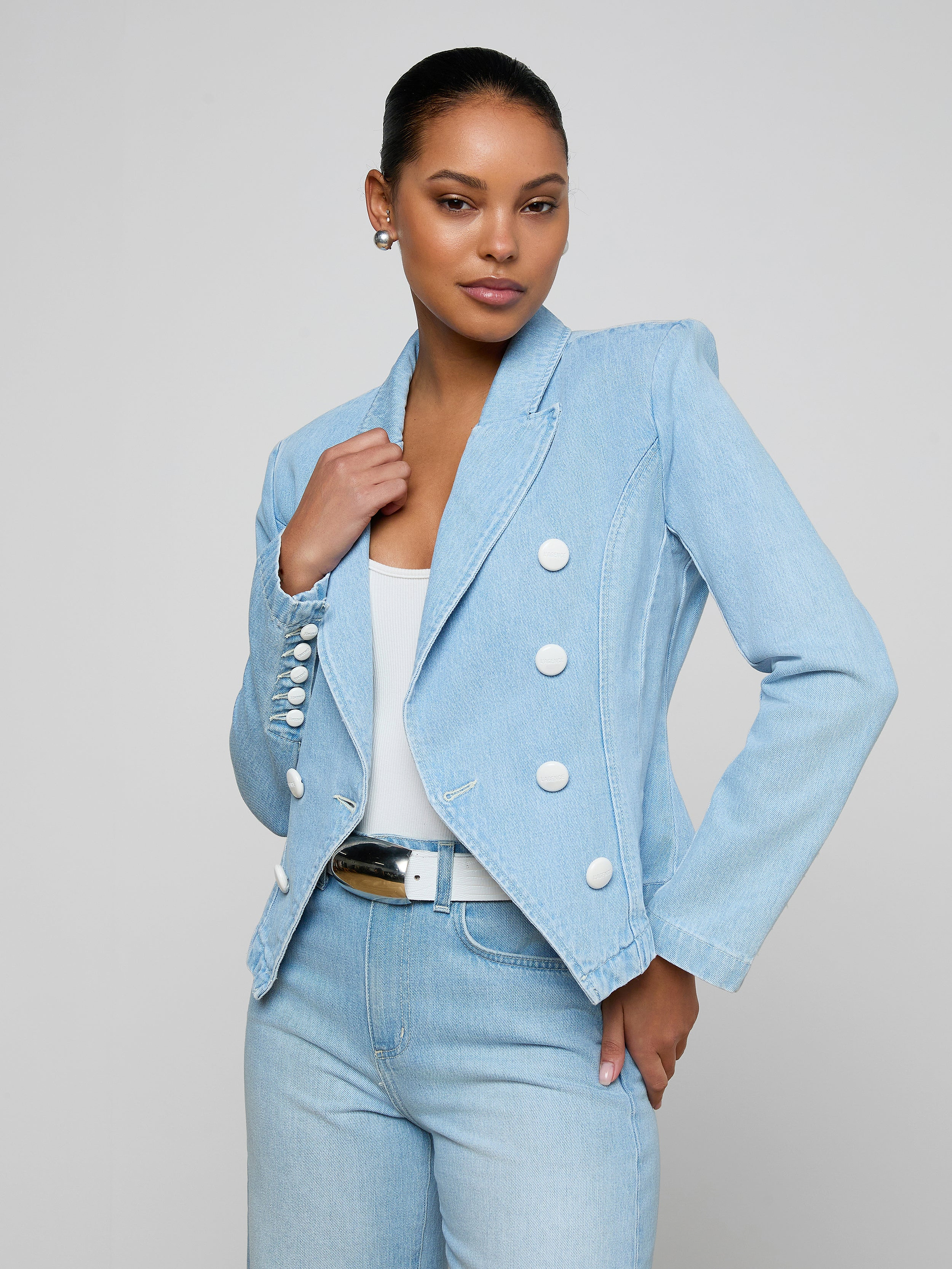 A woman stands confidently against a plain light background, wearing the Emerson Structured Blazer—a light wash, all-cotton denim double-breasted blazer with white buttons—paired with a white top, light blue jeans, and a black belt.