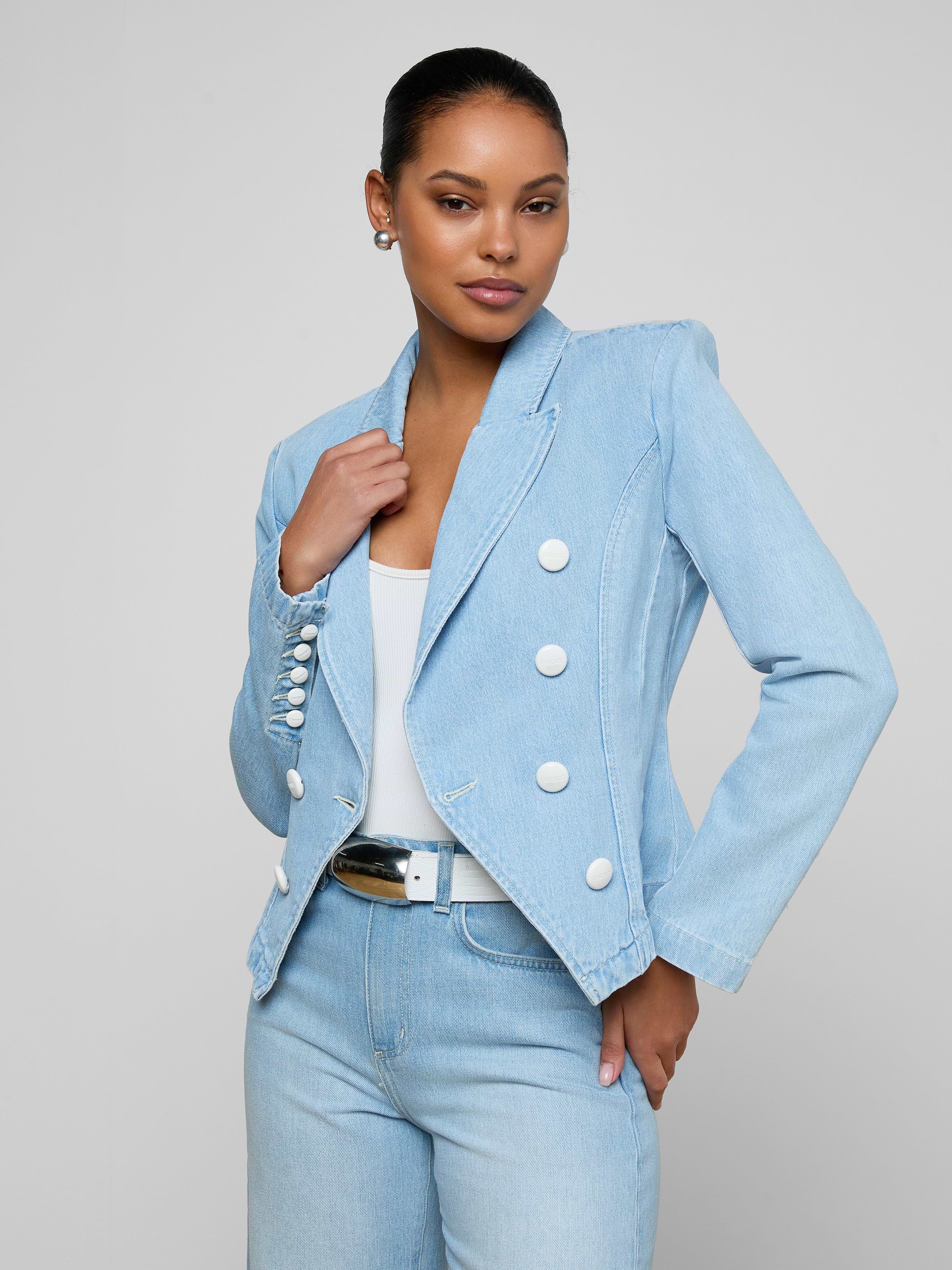 A woman stands confidently against a plain light background, wearing the Emerson Structured Blazer—a light wash, all-cotton denim double-breasted blazer with white buttons—paired with a white top, light blue jeans, and a black belt.