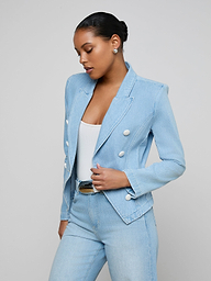 A woman wears the Emerson Structured Blazer—a light wash, double-breasted style with white buttons—over a white top and matching light blue, all-cotton denim jeans. She stands against a plain light background, looking down with her hair in a neat bun.