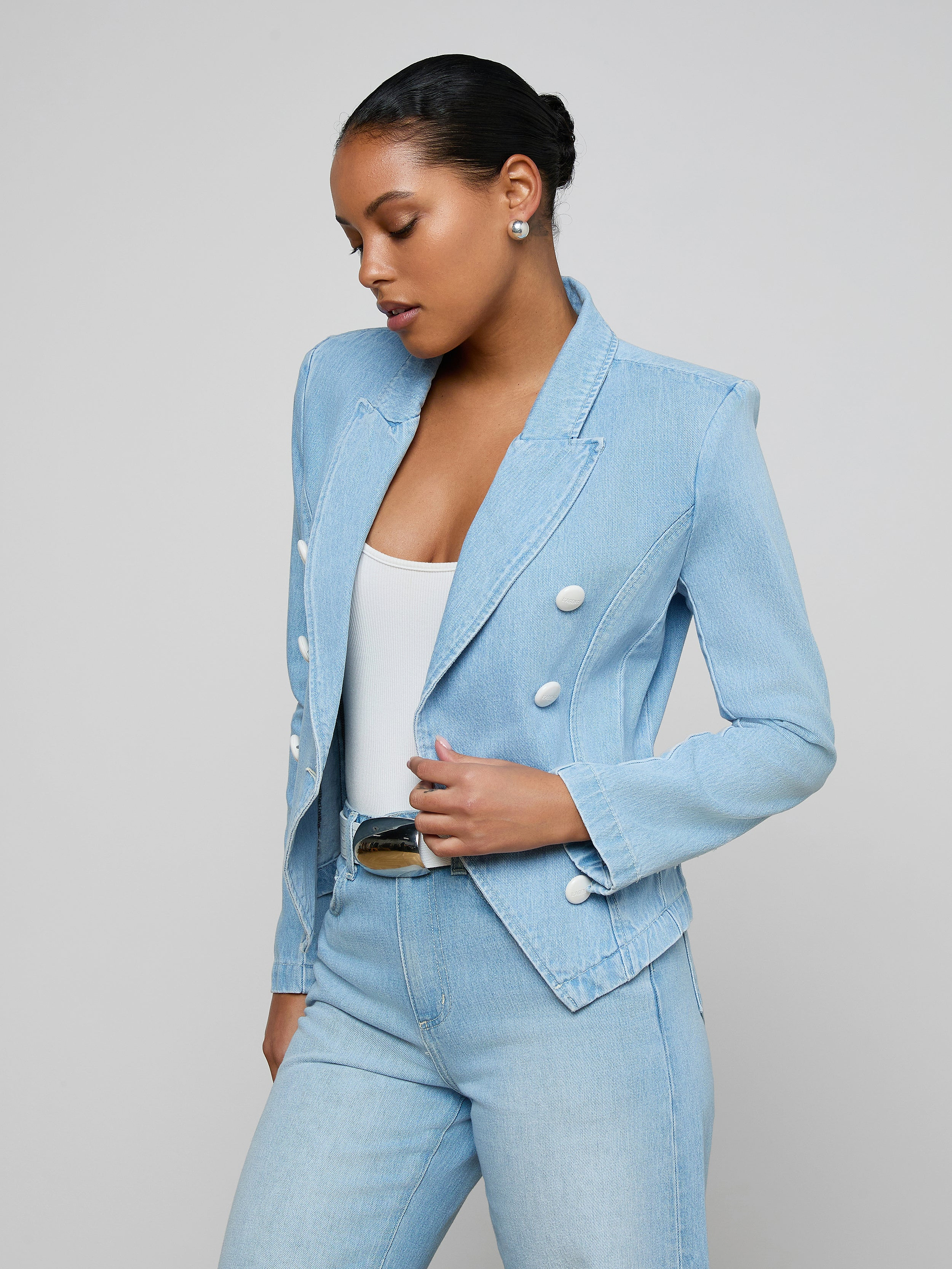 A woman wears the Emerson Structured Blazer—a light wash, double-breasted style with white buttons—over a white top and matching light blue, all-cotton denim jeans. She stands against a plain light background, looking down with her hair in a neat bun.