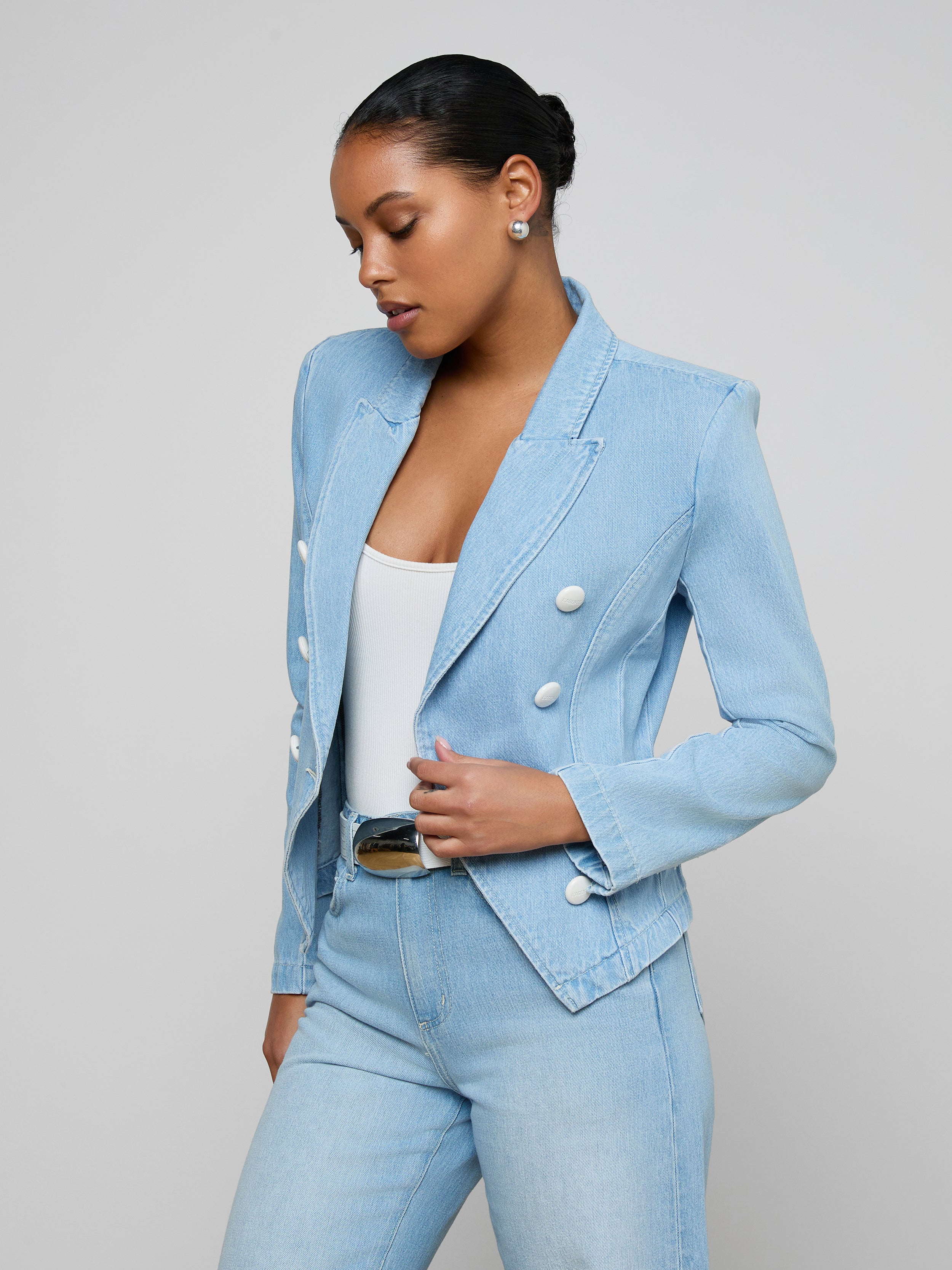 A woman wears the Emerson Structured Blazer—a light wash, double-breasted style with white buttons—over a white top and matching light blue, all-cotton denim jeans. She stands against a plain light background, looking down with her hair in a neat bun.
