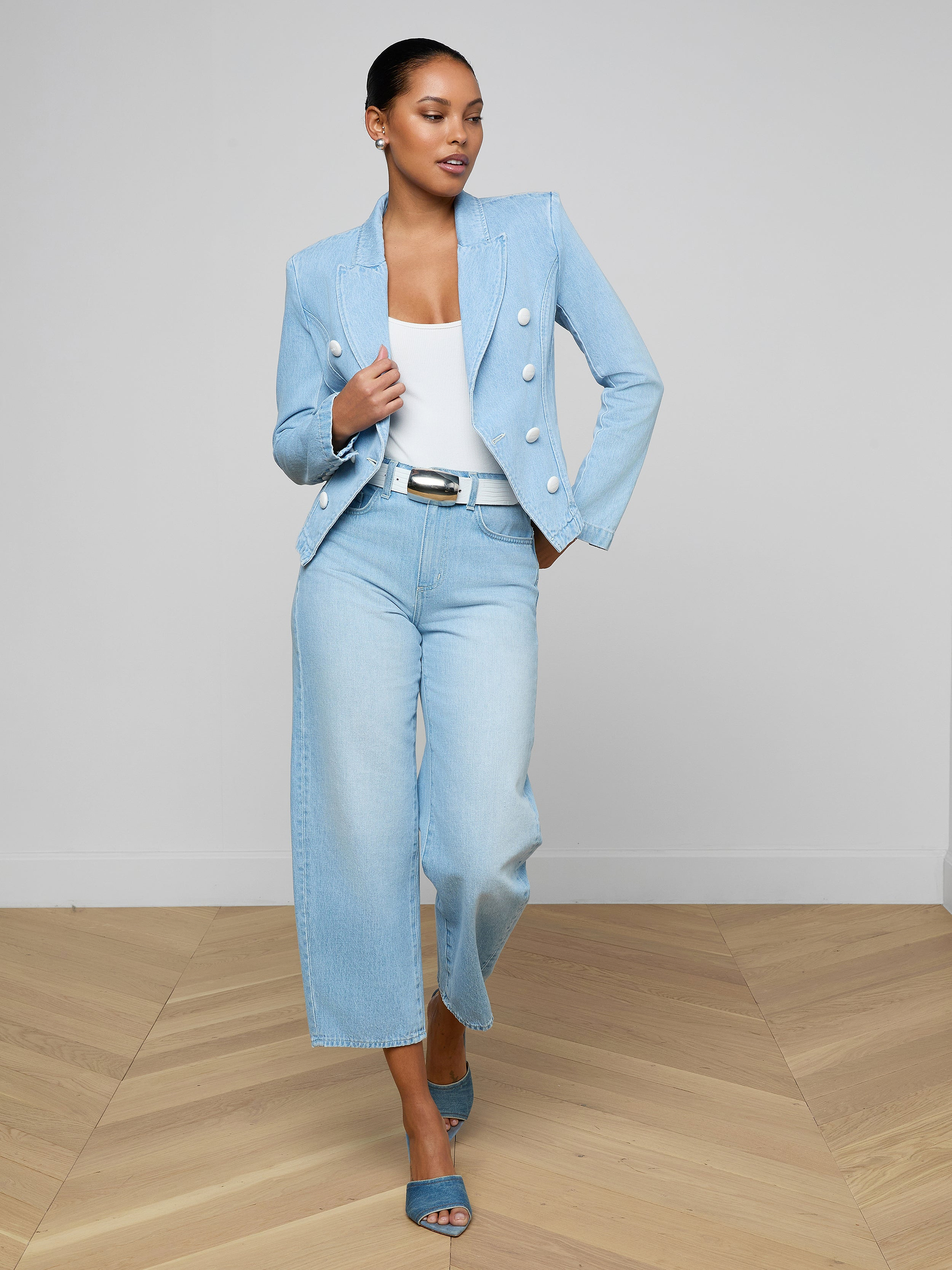 A woman wearing the Emerson Structured Blazer in light blue, paired with a white top and wide-legged, light-wash all-cotton denim jeans with blue heels, stands confidently against a plain white background.