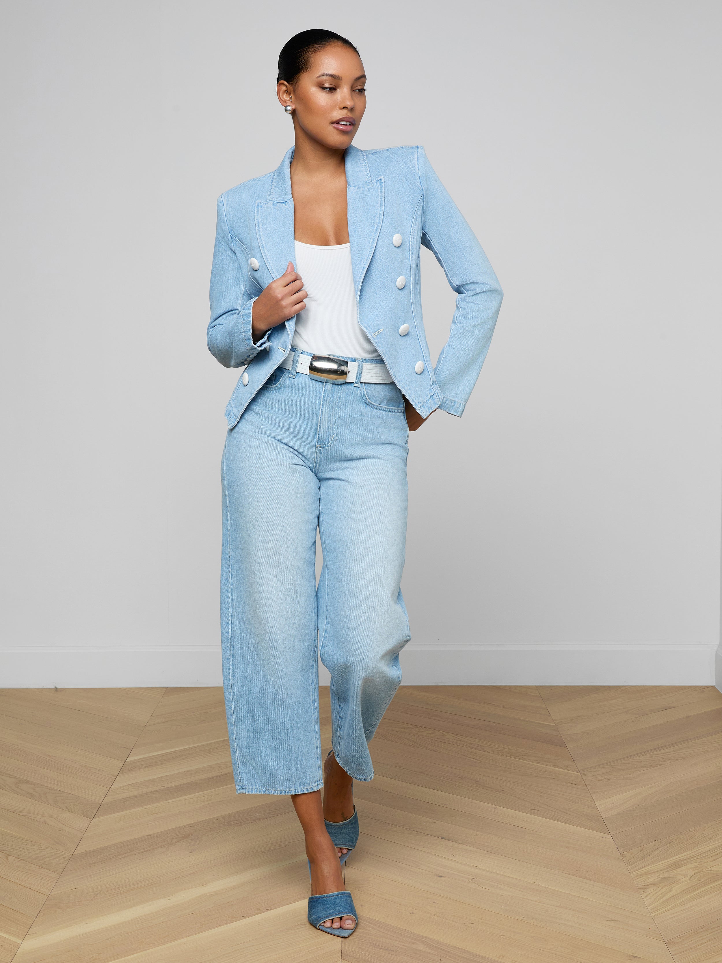 A woman wearing the Emerson Structured Blazer in light blue, paired with a white top and wide-legged, light-wash all-cotton denim jeans with blue heels, stands confidently against a plain white background.