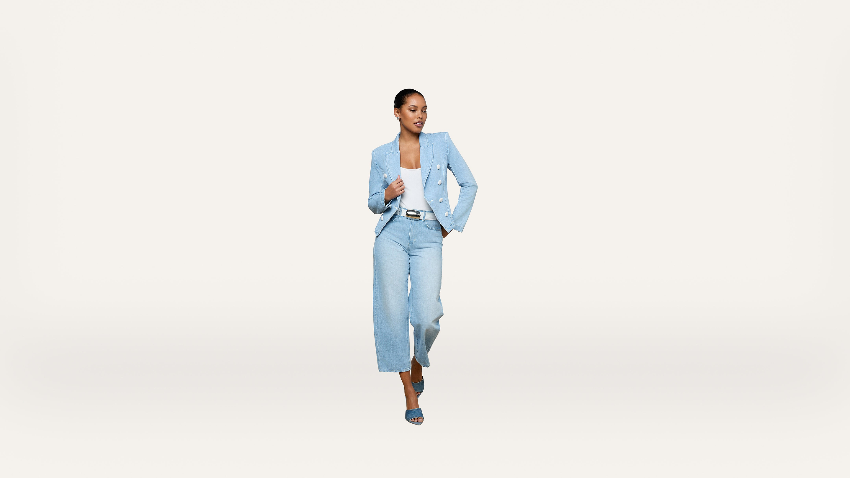 Woman in a light blue pantsuit and heels walking confidently against a plain white background.