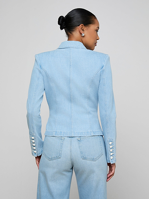 A person stands with their back turned, wearing the Emerson Structured Blazer in light blue denim with decorative sleeve buttons and matching high-waisted jeans. Their hair is in a neat bun against a plain, light-colored background.