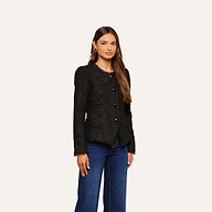 A woman with long brown hair wears the Nerina Tweed Blazer, a collarless black jacket, paired with dark blue jeans as she stands against a plain light background.