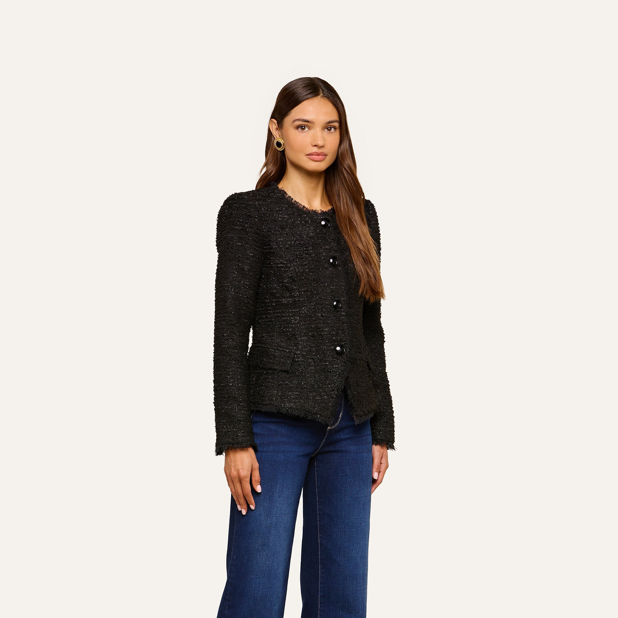 A woman with long brown hair wears the Nerina Tweed Blazer, a collarless black jacket, paired with dark blue jeans as she stands against a plain light background.