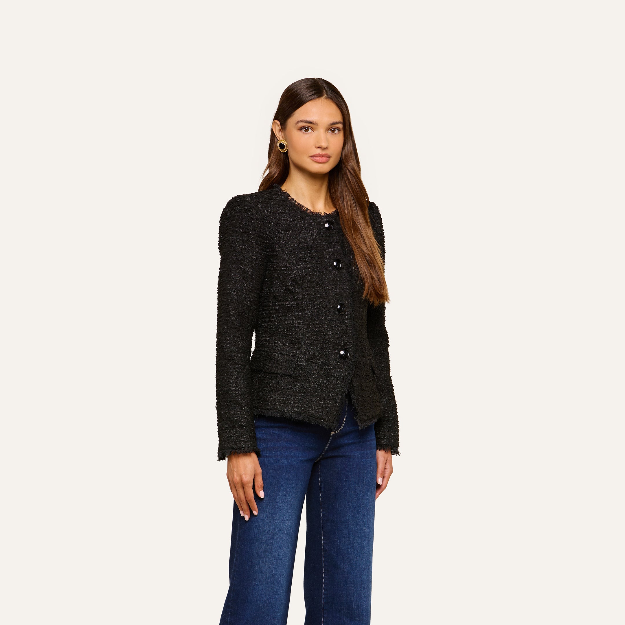 A woman with long brown hair wears the Nerina Tweed Blazer, a collarless black jacket, paired with dark blue jeans as she stands against a plain light background.