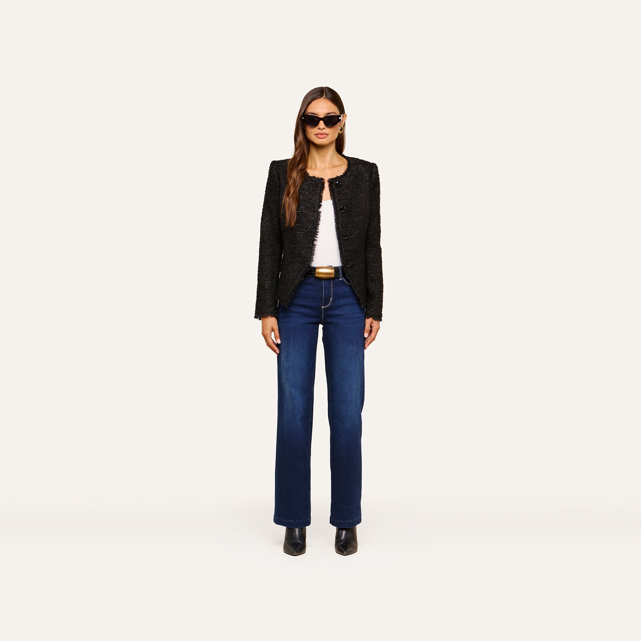 A woman stands facing forward, wearing sunglasses, the Nerina Tweed Blazer in black, a white top, dark blue jeans, and black shoes against a plain light-colored background.