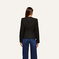 A woman stands facing away, wearing the Nerina Tweed Blazer—a collarless black tweed blazer—paired with blue jeans featuring white stitching, set against a plain light background.