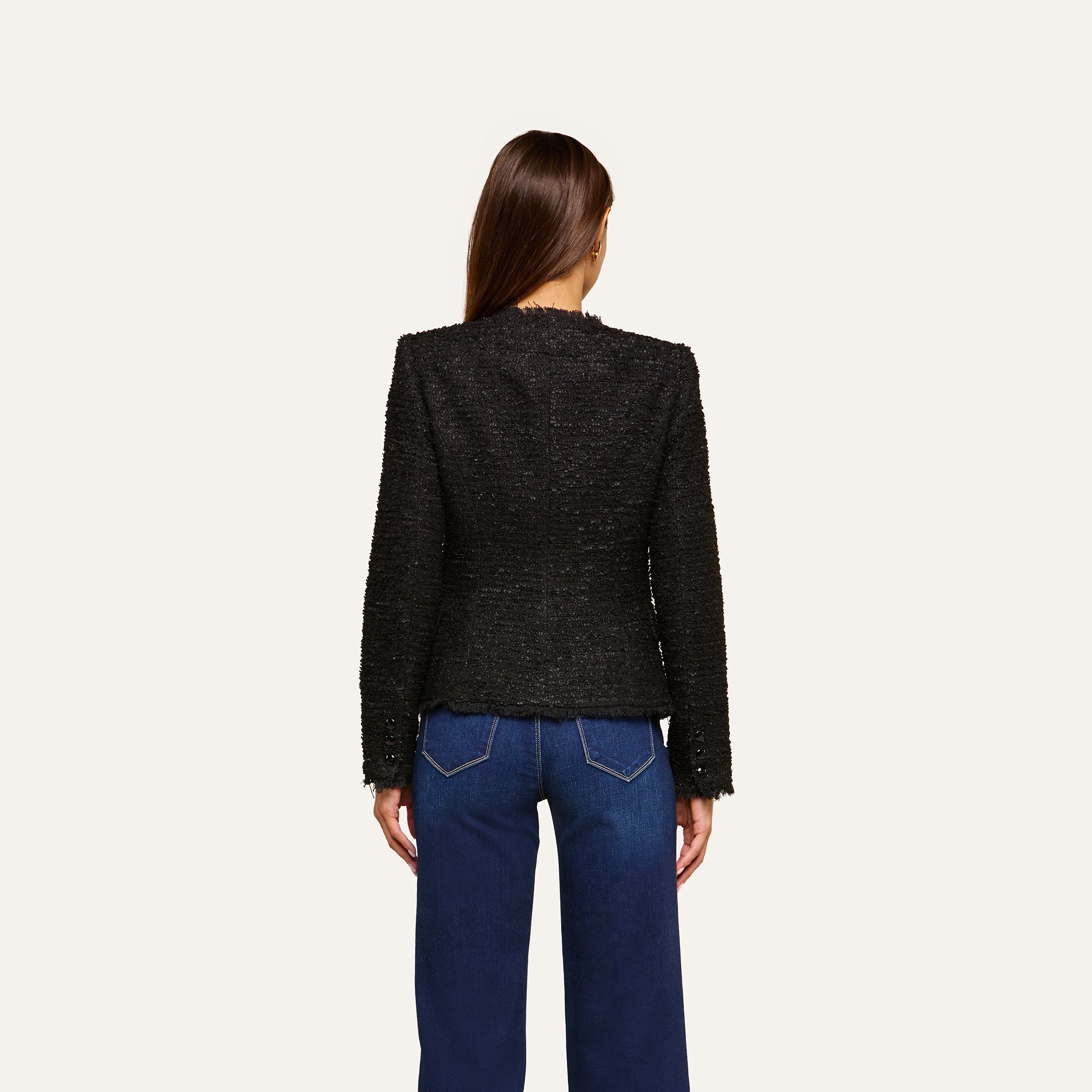 A woman stands facing away, wearing the Nerina Tweed Blazer—a collarless black tweed blazer—paired with blue jeans featuring white stitching, set against a plain light background.