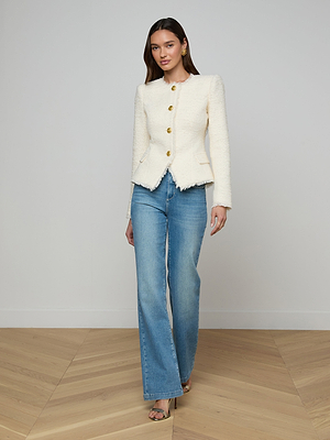 A woman stands indoors on a wooden floor, wearing the Nerina Tweed Blazer—a cream collarless blazer with gold buttons—styled with blue high-waisted jeans and open-toe heels. Her long brown hair is loose, and she faces the camera with a slight smile.