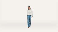 A woman wearing a white blazer and blue jeans walks forward against a plain white background.