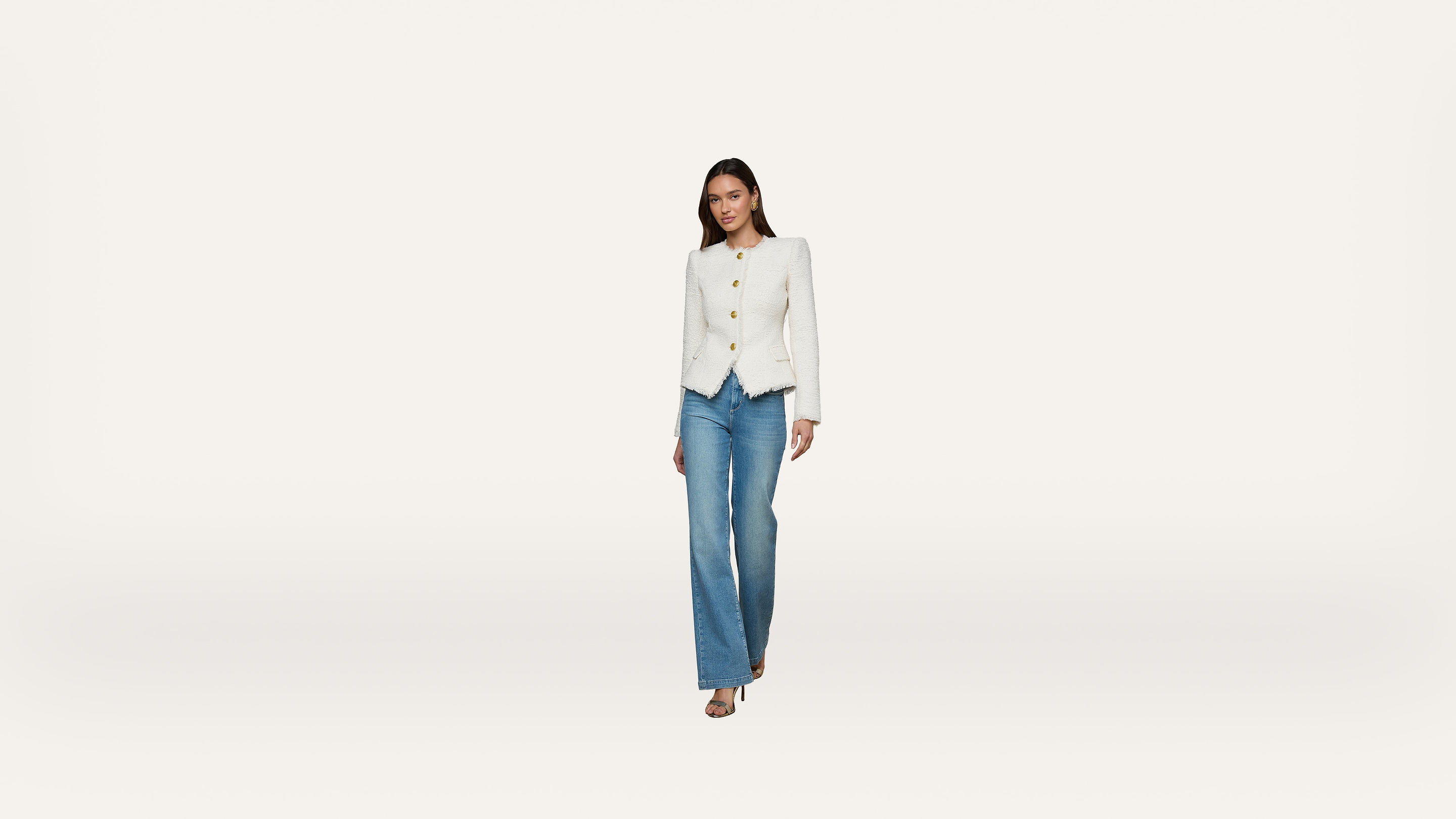 A woman wearing a white blazer and blue jeans walks forward against a plain white background.