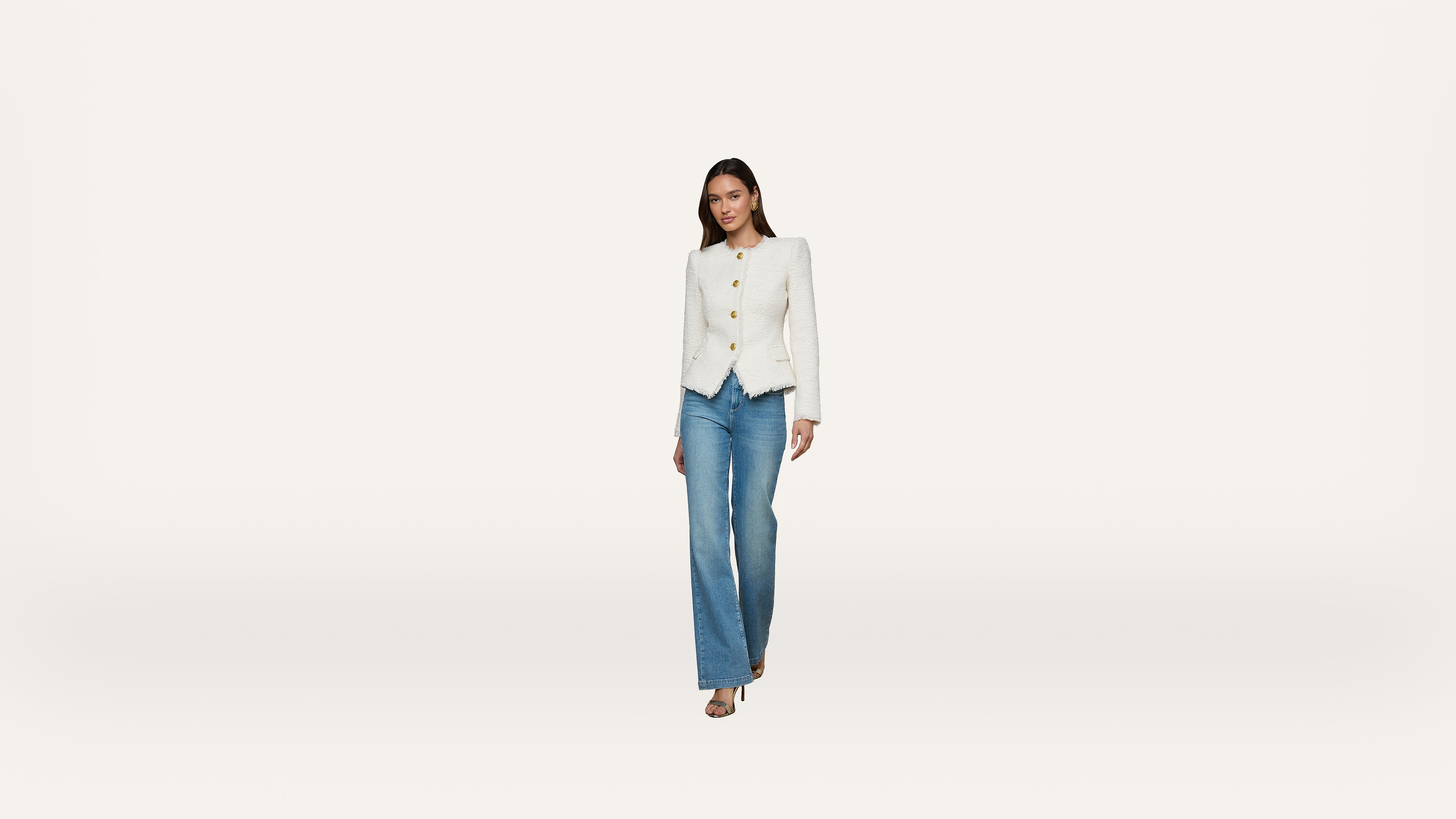 A woman wearing a white blazer and blue jeans walks forward against a plain white background.