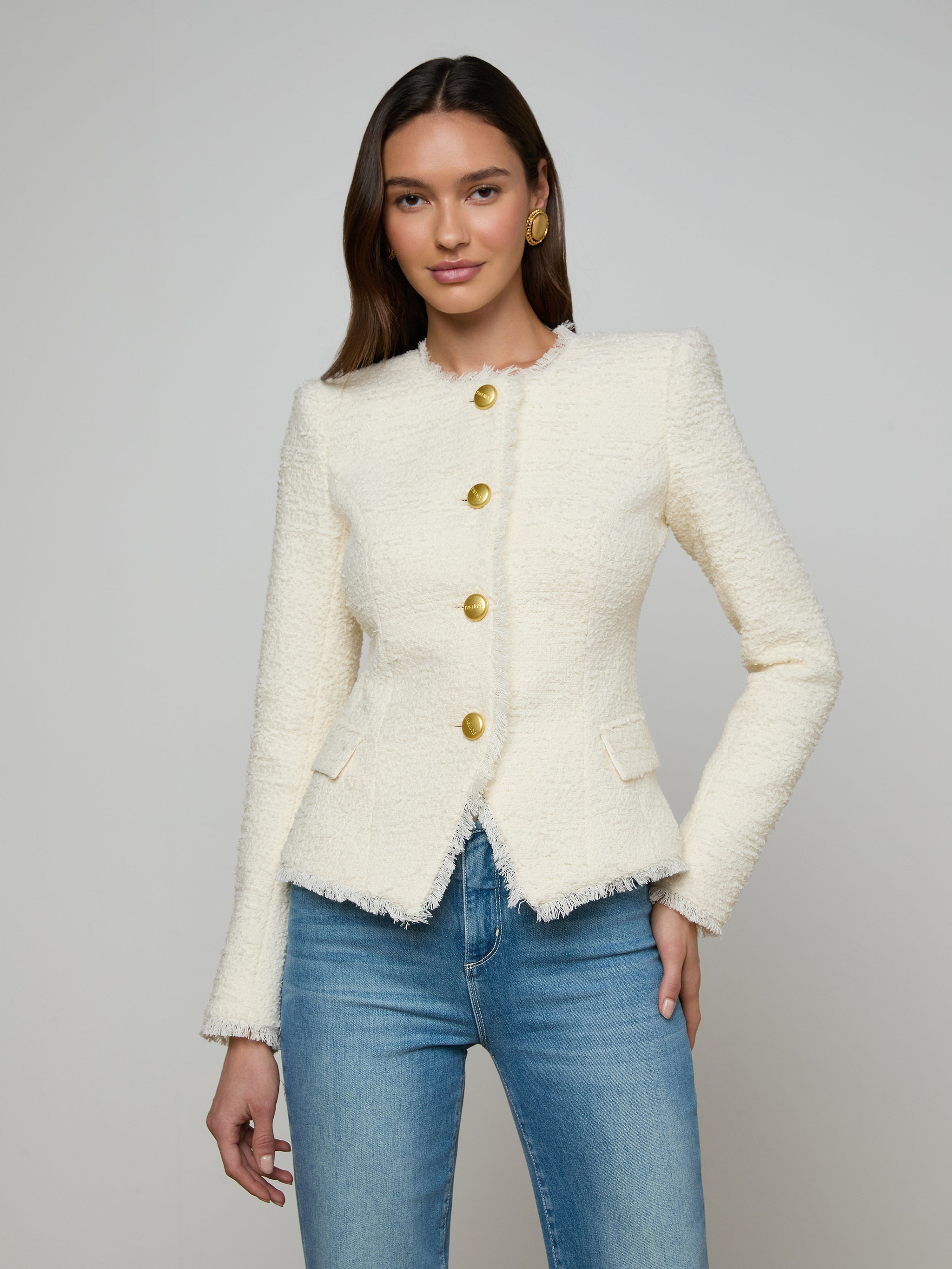 A woman wears the Nerina Tweed Blazer—a fitted, textured, white collarless jacket with gold buttons—paired with blue jeans, standing against a plain light background.