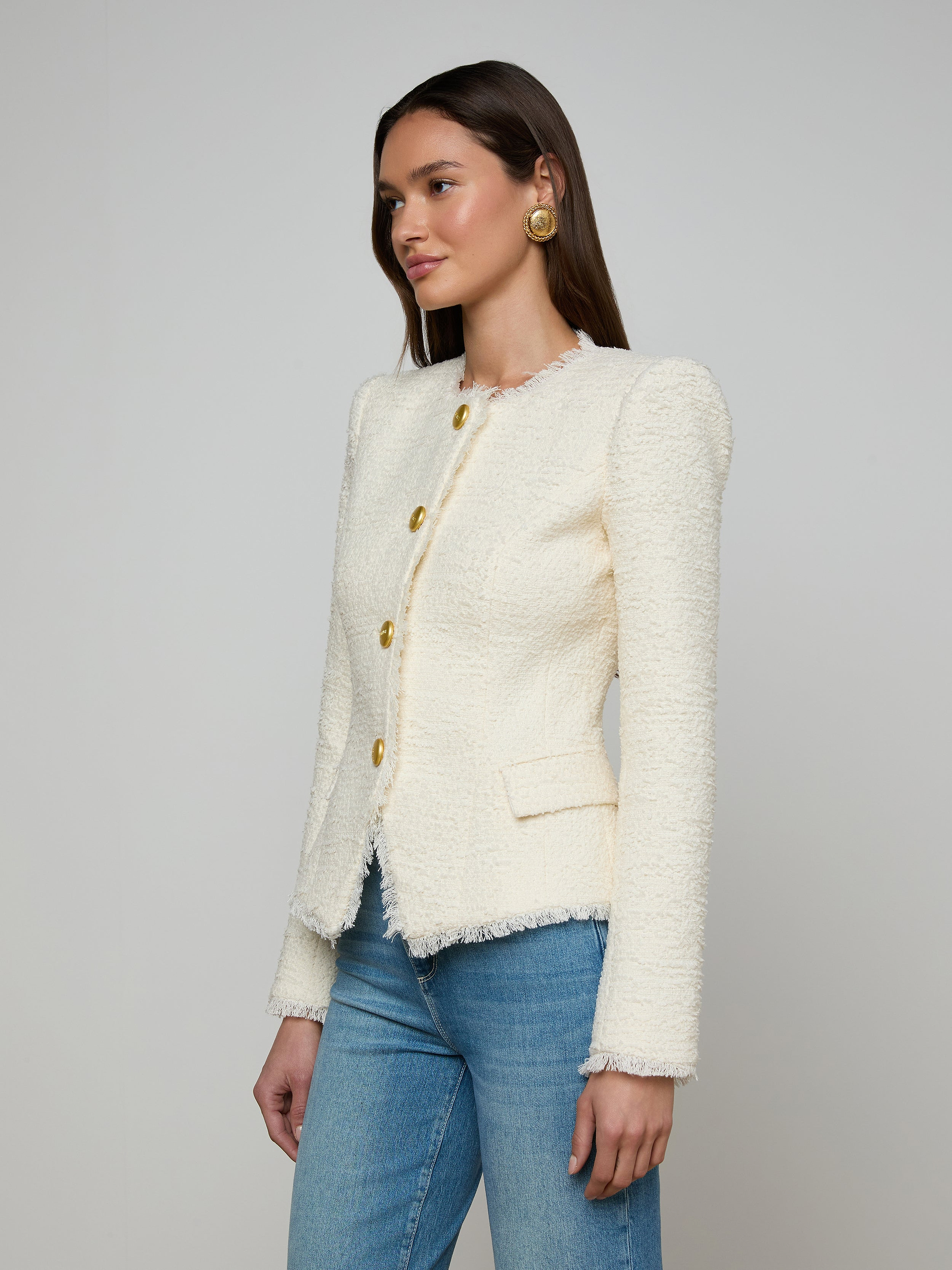 A woman wearing the Nerina Tweed Blazer—featuring a white textured finish and gold buttons—paired with blue jeans, stands against a plain light background.