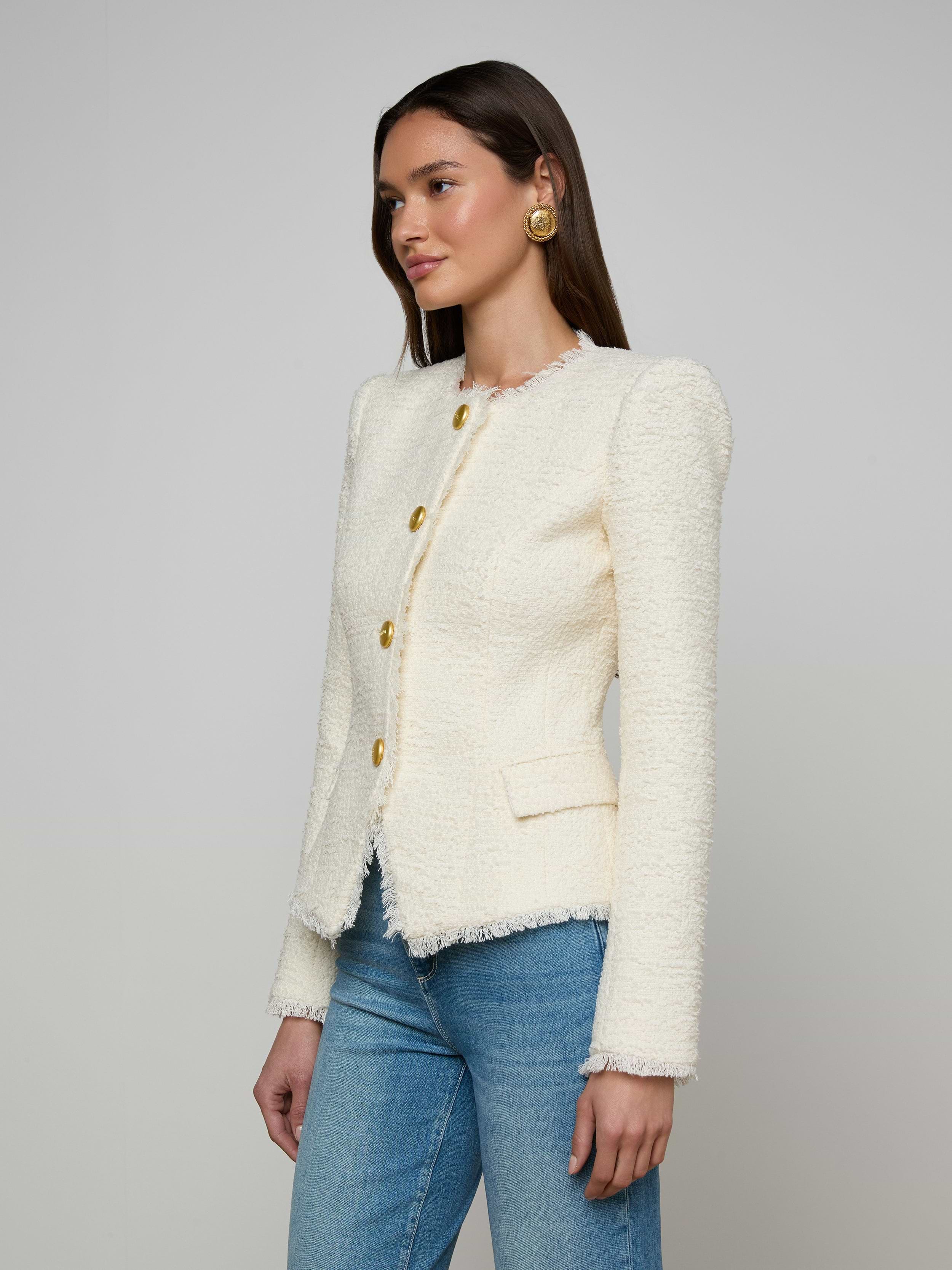 A woman wearing the Nerina Tweed Blazer—featuring a white textured finish and gold buttons—paired with blue jeans, stands against a plain light background.