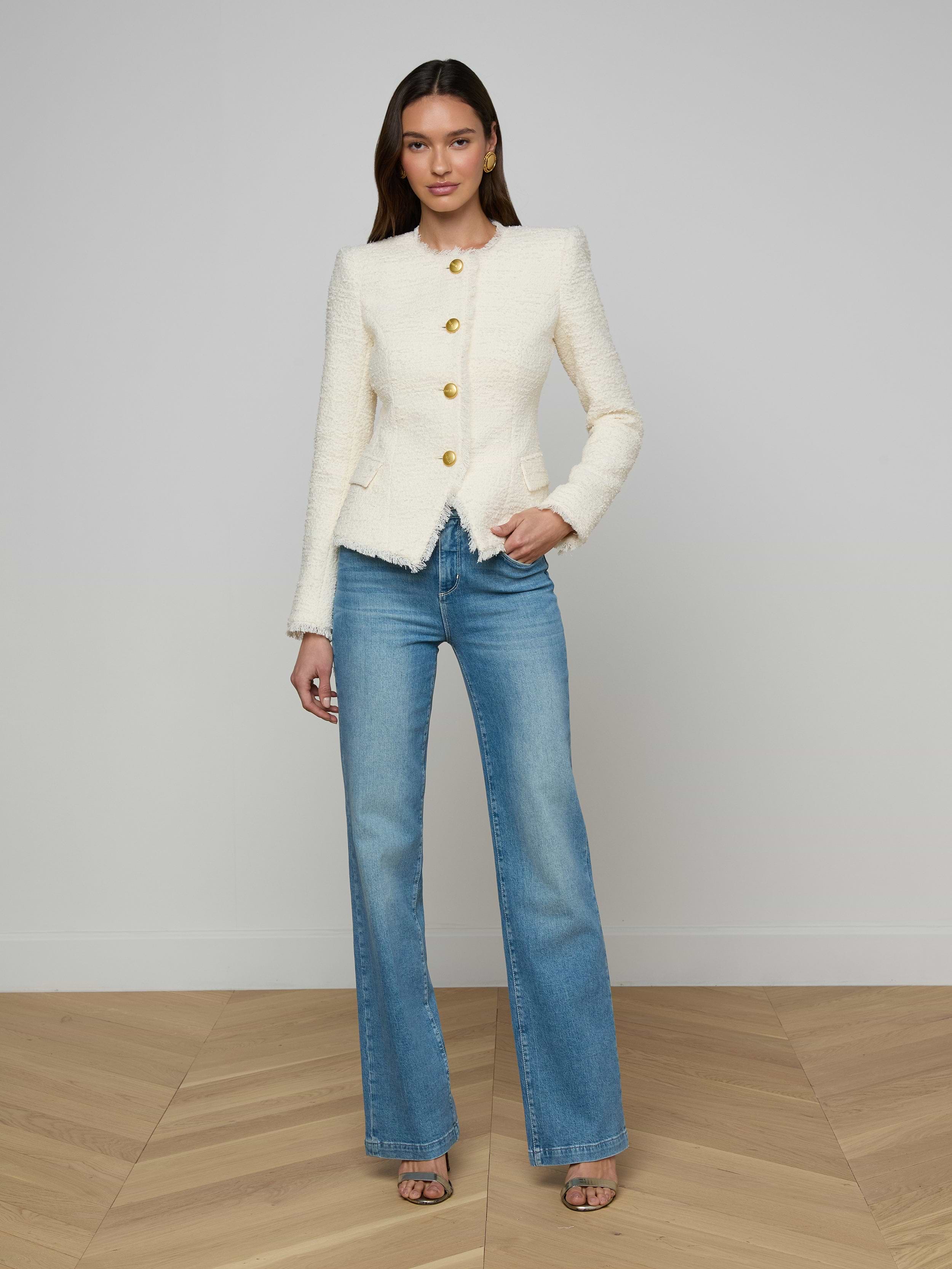 A woman stands against a plain white background, wearing the Nerina Tweed Blazer styled as a statement piece, paired with blue jeans and open-toe heels. She has long hair and one hand in her pocket.