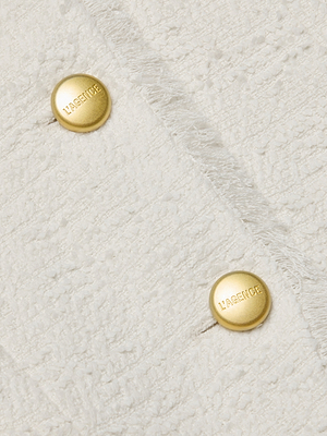 Close-up of textured cream-colored fabric from the Nerina Tweed Blazer. Two gold buttons embossed with “L’AGENCE” stand out on the soft, slightly fuzzy collarless jacket, highlighting its visible seams.