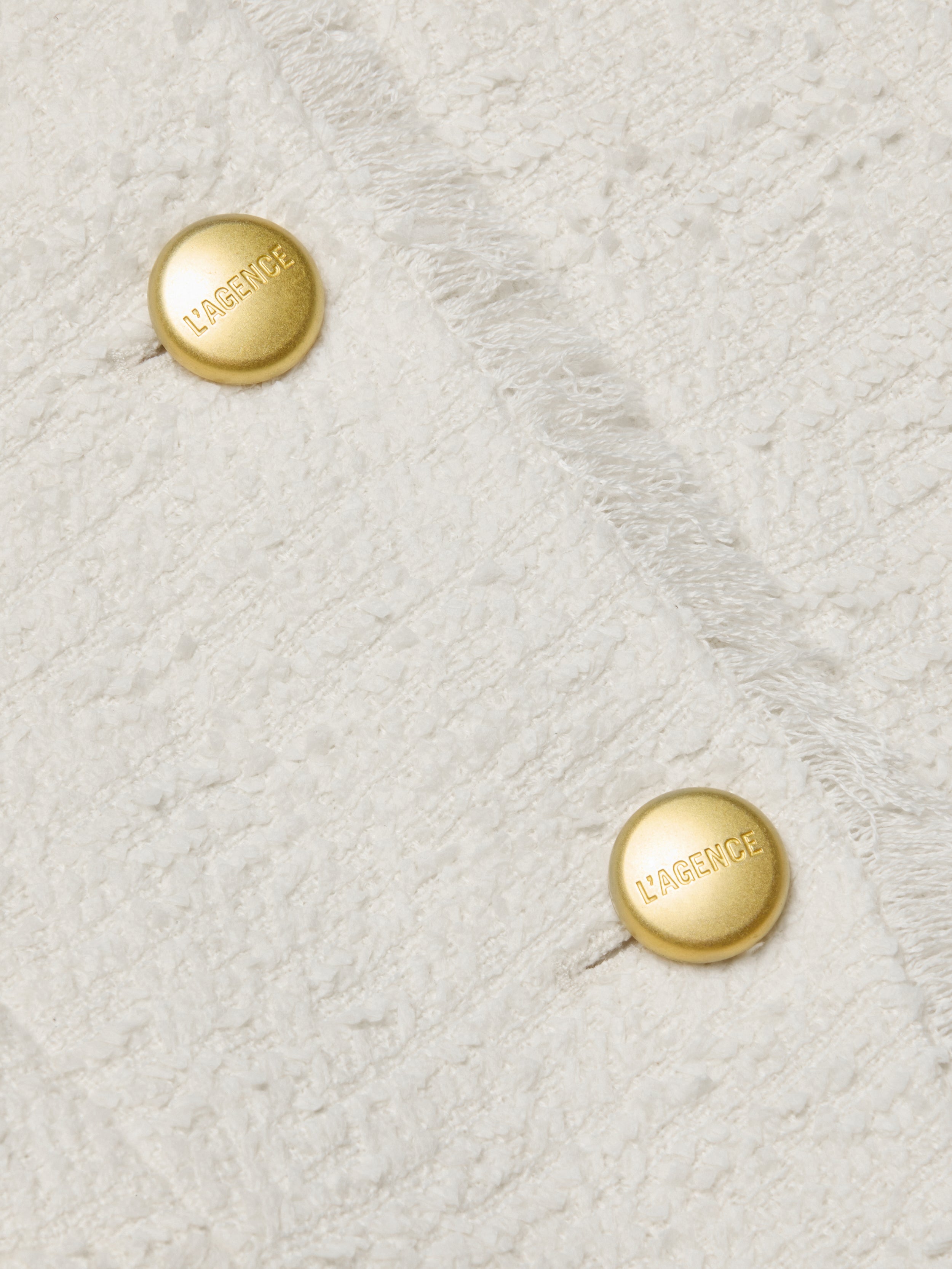 Close-up of the textured white fabric from the Nerina Tweed Blazer, featuring a collarless neckline and two parallel gold buttons engraved with LAGENCE. The fabric showcases subtle frayed edges for added detail.
