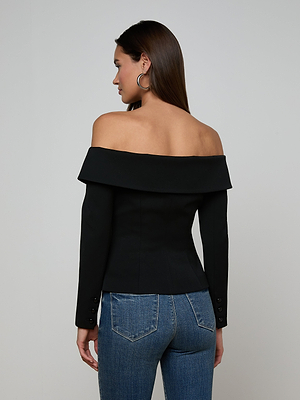 A woman with long brown hair wears the Ingrid Blazer Top—an off-the-shoulder black piece with a folded neckline and long sleeves—paired with blue jeans. She stands facing away, showcasing the back against a plain light background.