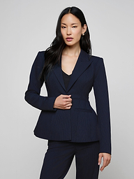 A woman with long black hair wears the sculptural Therese Blazer in navy pinstripe and stands against a plain light background, facing forward with one hand touching her jacket.