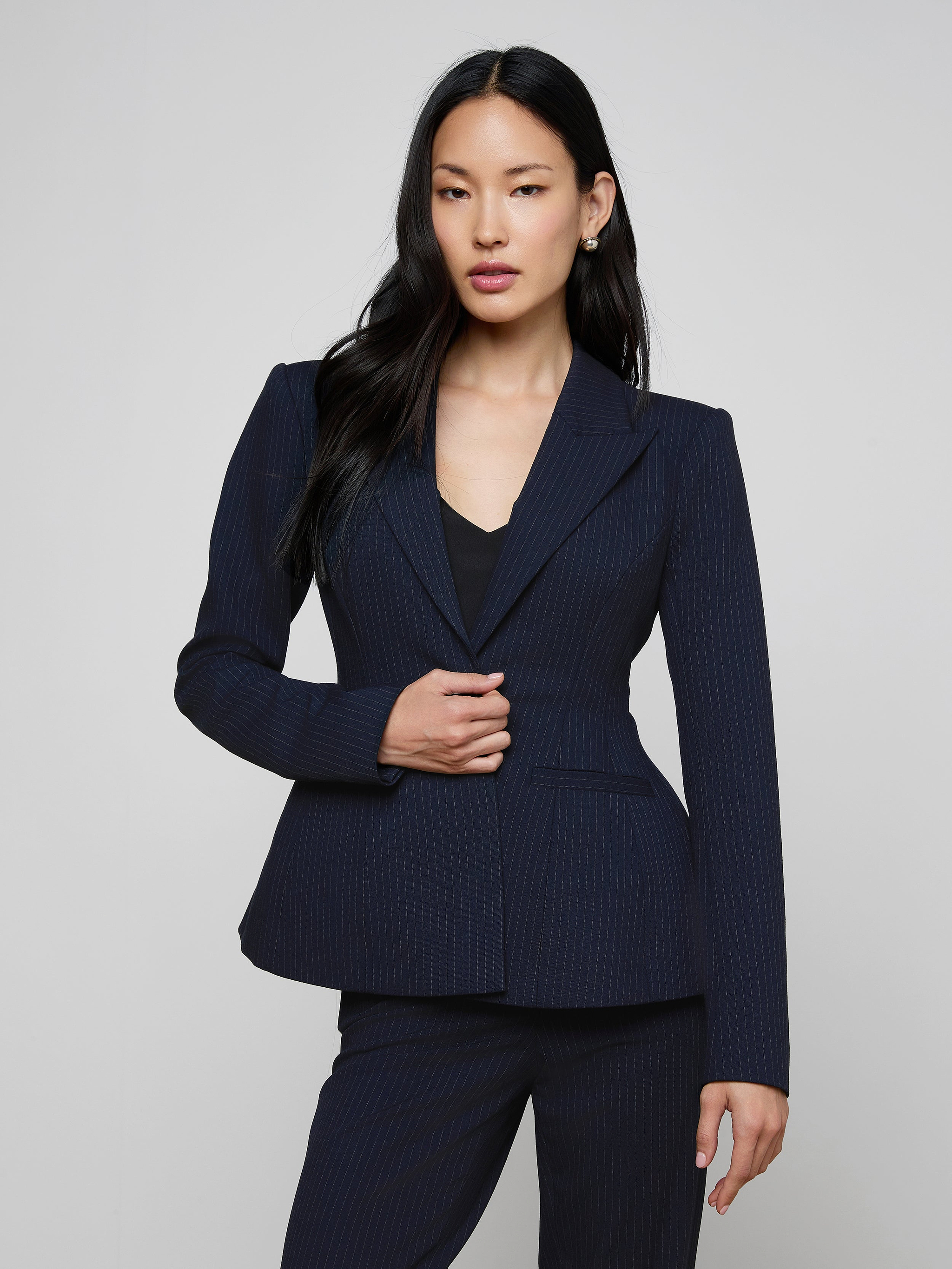 A woman with long black hair wears the sculptural Therese Blazer in navy pinstripe and stands against a plain light background, facing forward with one hand touching her jacket.