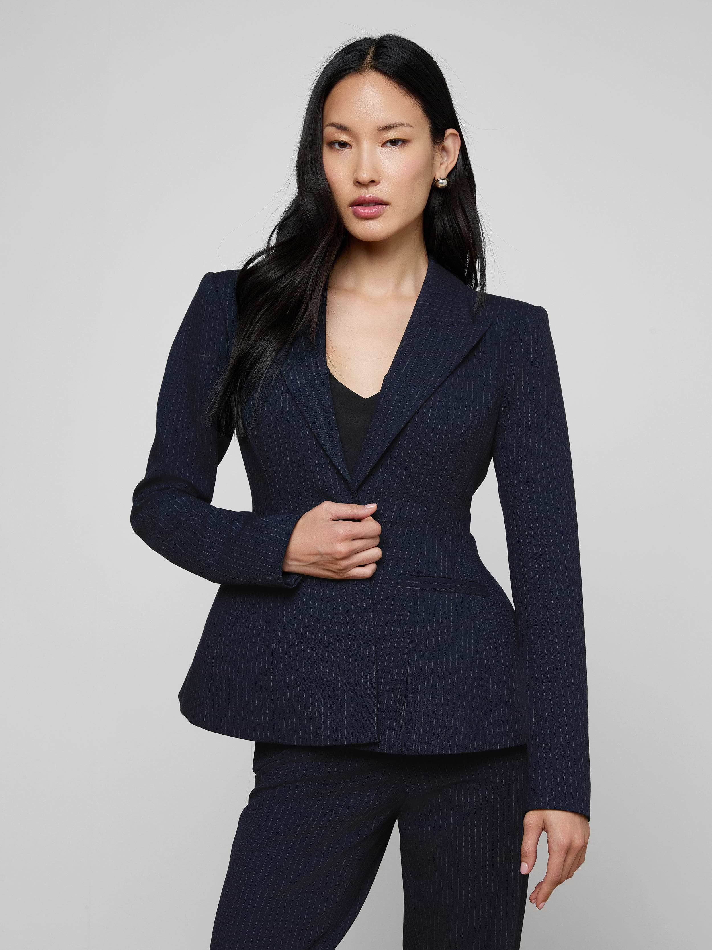 A woman with long black hair wears the sculptural Therese Blazer in navy pinstripe and stands against a plain light background, facing forward with one hand touching her jacket.