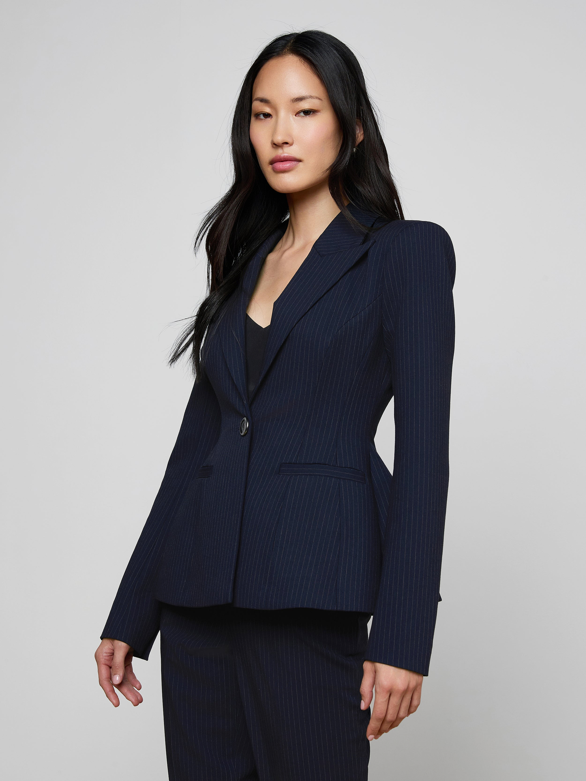 A woman with long dark hair wears the fitted navy pinstripe Therese Blazer and poses confidently against a plain light background.