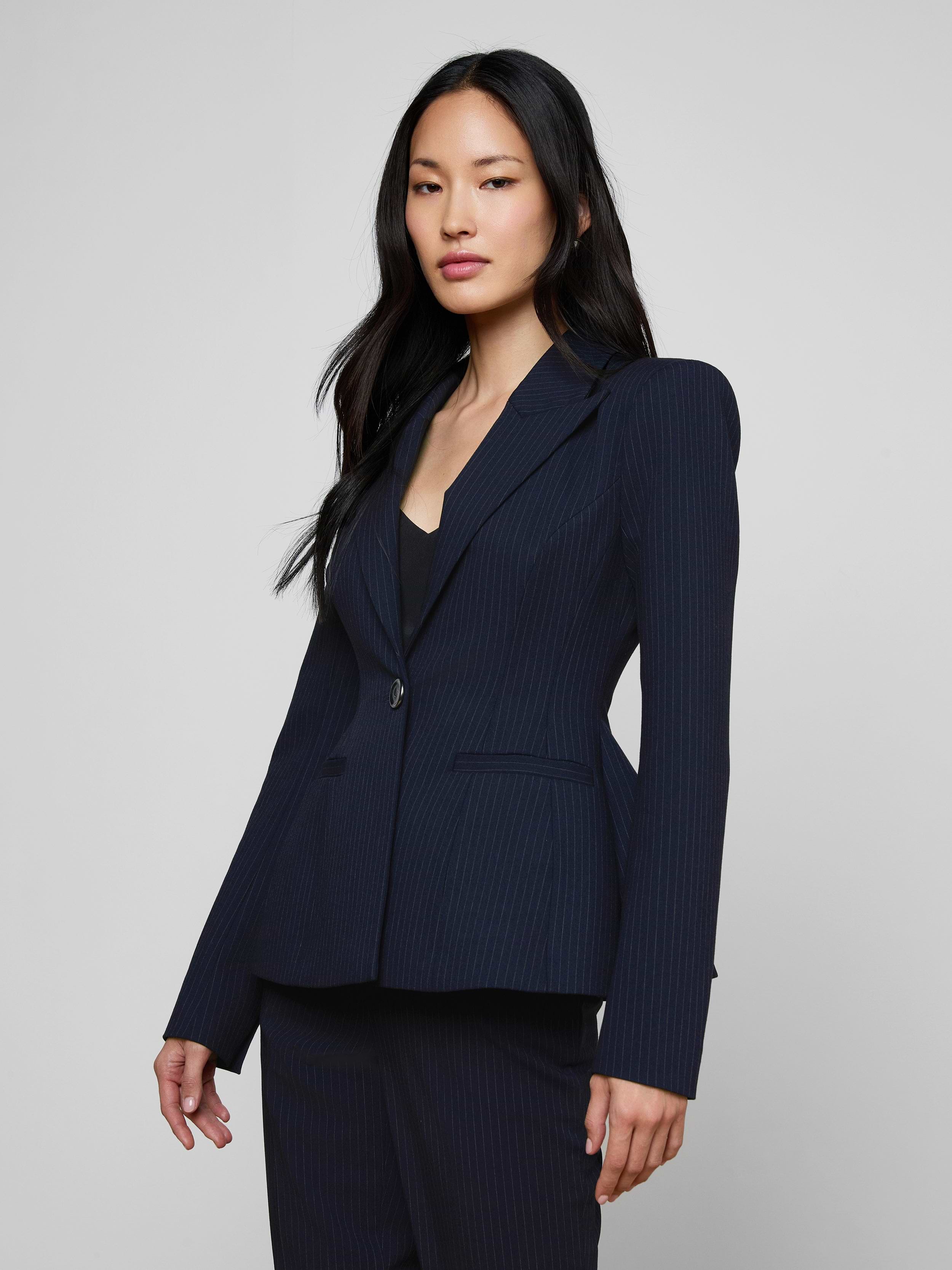 A woman with long dark hair wears the fitted navy pinstripe Therese Blazer and poses confidently against a plain light background.