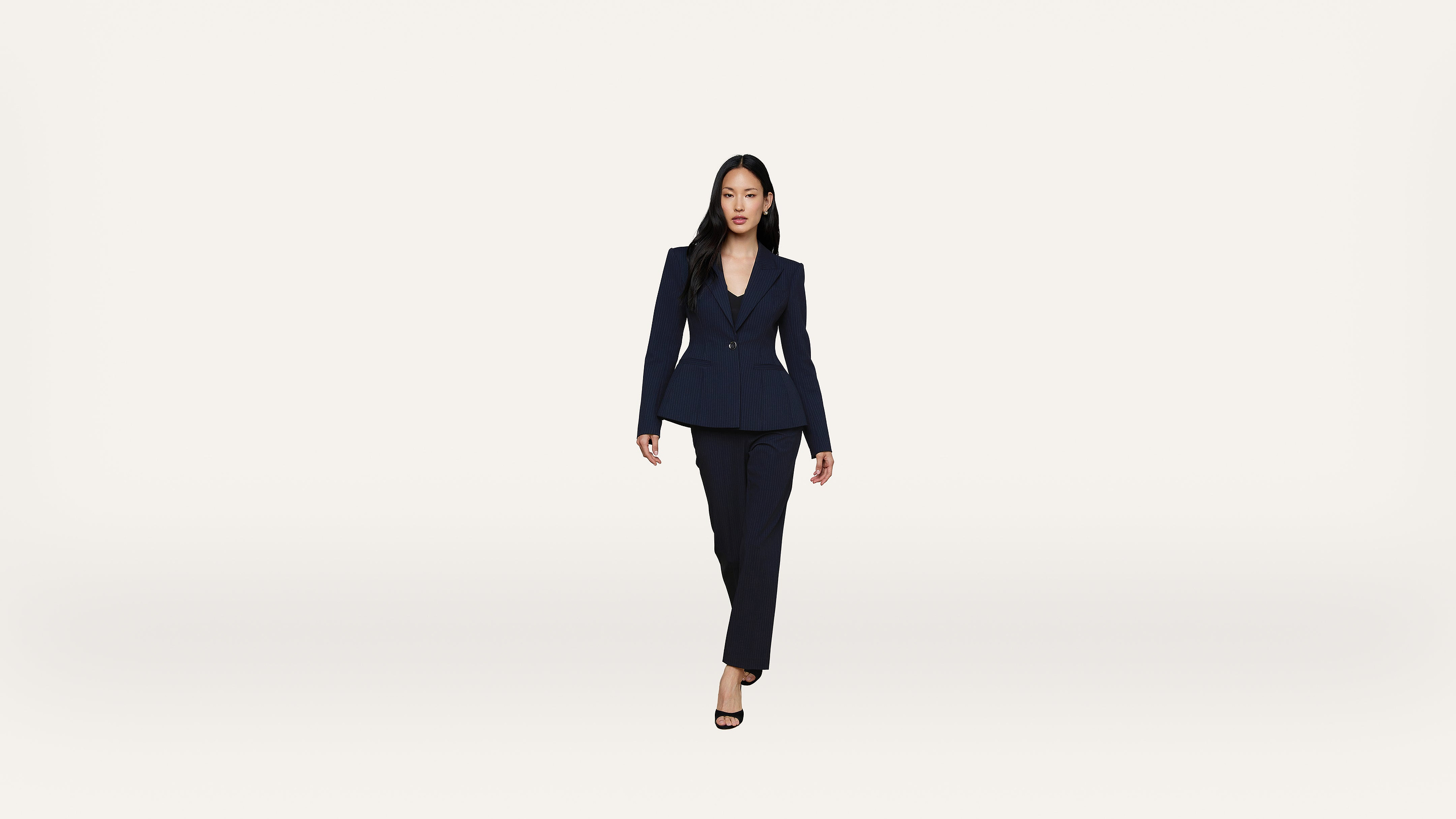 A woman in a dark suit and heels walks confidently toward the camera against a plain white background.