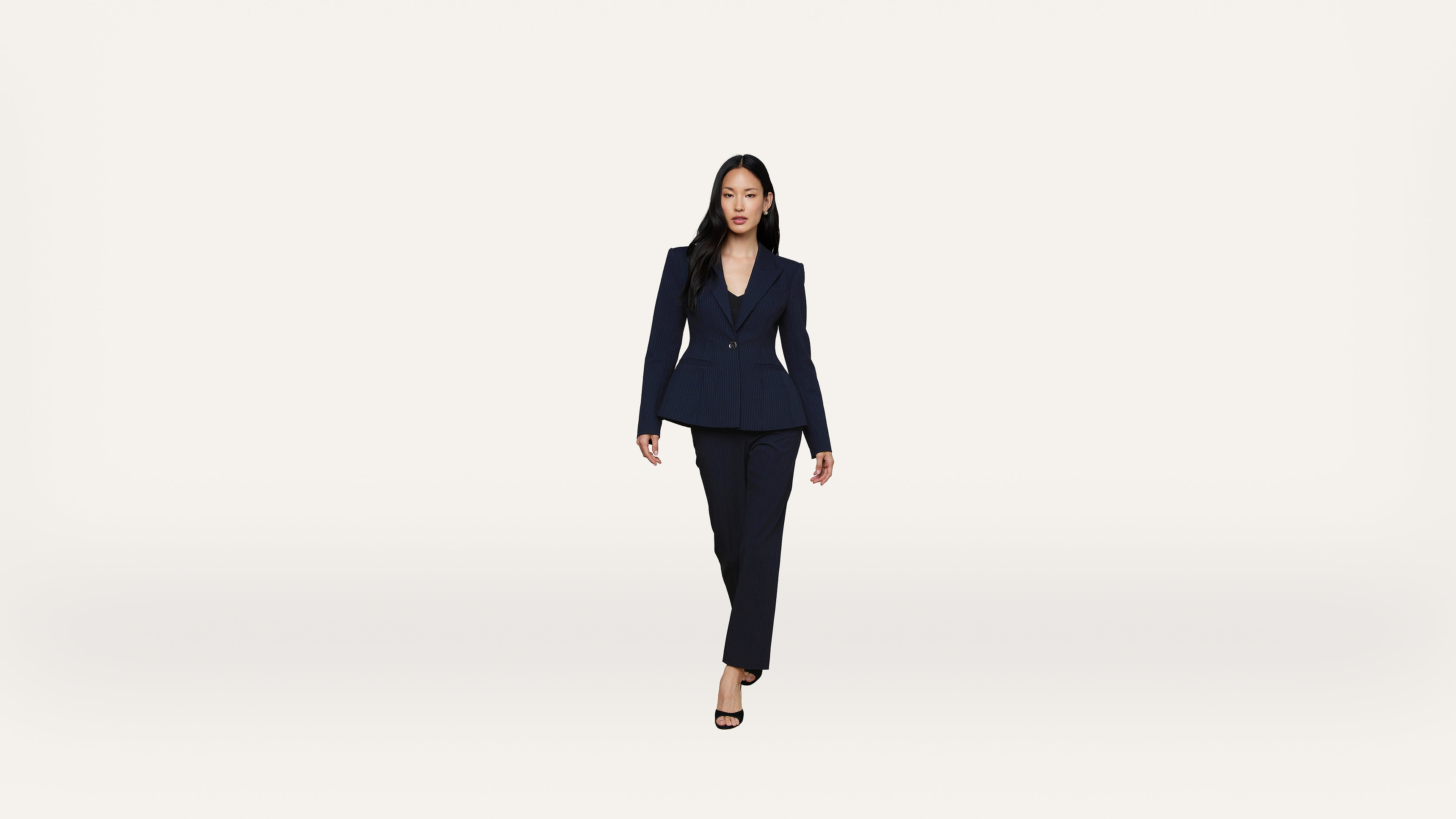 A woman in a dark suit and heels walks confidently toward the camera against a plain white background.