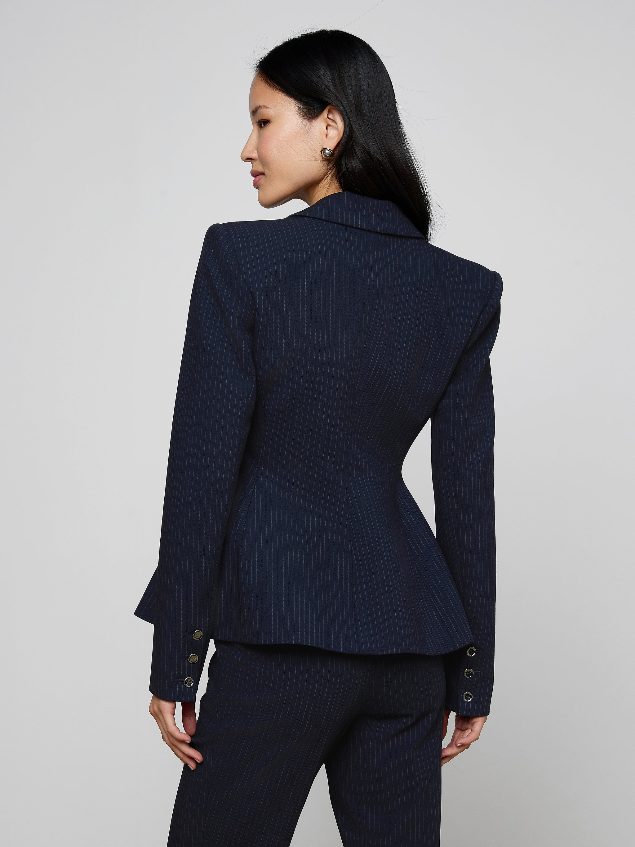 A woman with long dark hair, wearing the Therese Blazer in navy pinstripe, stands facing away and slightly to the side against a light background.