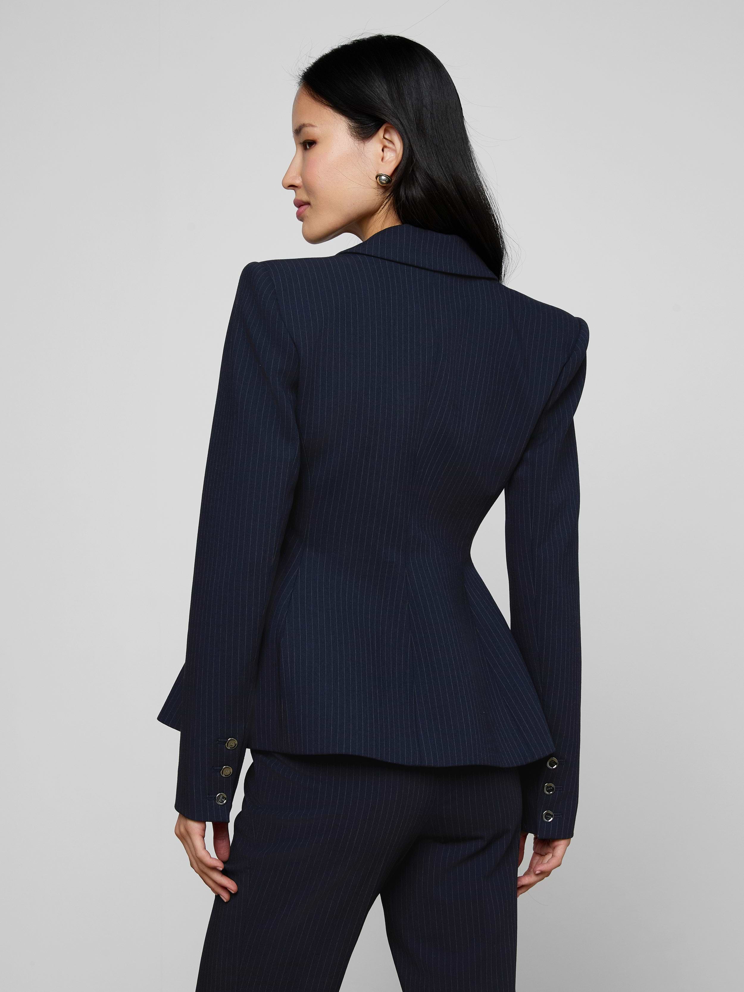 A woman with long dark hair, wearing the Therese Blazer in navy pinstripe, stands facing away and slightly to the side against a light background.