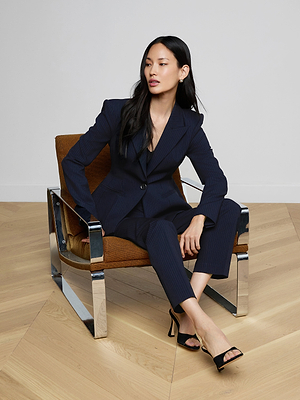 A woman in the Therese Blazer with navy pinstripes and black heels sits cross-legged on a brown chair with metal arms, looking to the side on a light wooden floor against a white wall.