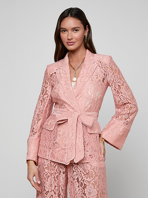 A woman with long brown hair wears the Jordana Lace Blazer—a pink sheer floral lace belted blazer and matching pants—standing against a light background. She styles it with layered necklaces and earrings for a chic Spring 2026 look.