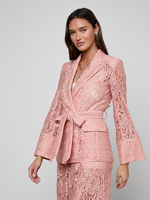 A woman in the Jordana Lace Blazer and matching pants, featuring pink sheer floral lace with a belt, stands confidently against a light gray background—an elegant look for Spring 2026.