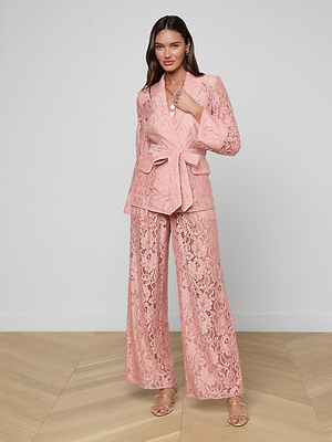 Indoors on a wooden floor, a woman wears the Jordana Lace Blazer—a sheer pink floral lace suit with a belted blazer and wide-leg pants—styled for Spring 2026. She pairs it with strappy heels and long hair, exuding calm confidence.